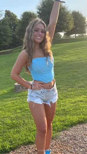 Woman in blue crop top and denim shorts posing outdoors on grassy hill.