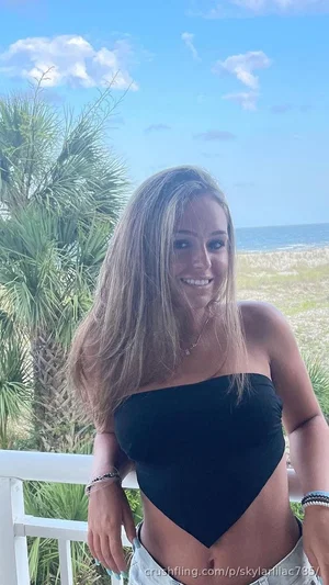 Woman with long blonde hair in black strapless top and light pants, standing outdoors near beach