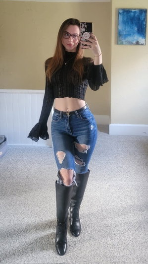Woman in black crop top, ripped jeans, and black boots takes mirror selfie indoors.