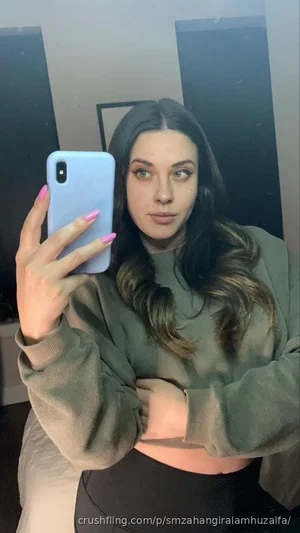 Woman with long dark hair in olive green crop top and black pants, taking mirror selfie indoors.