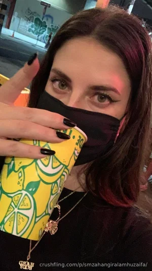 Woman with dark hair and black mask holding a yellow lemon cup, wearing a black top and necklace.