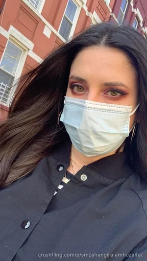 Woman with long dark hair wearing a face mask and black jacket outdoors in front of a brick