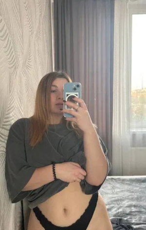 Brown-haired woman in dark top taking bedroom mirror selfie.