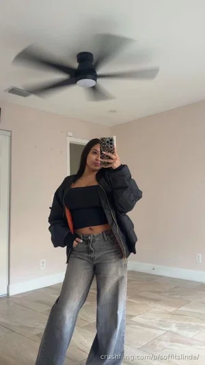 Woman in black crop top and puffer jacket, gray jeans, taking mirror selfie in room with ceiling