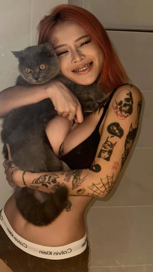 Red-haired woman with tattoos holding a gray cat indoors.
