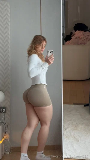 Woman in white long-sleeve top and beige shorts taking mirror selfie in bedroom.