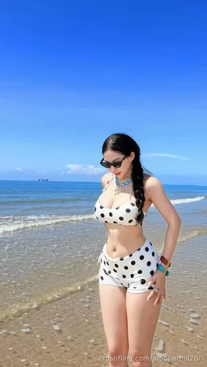Woman in polka-dot bikini standing on beach, wearing sunglasses and turquoise necklace.