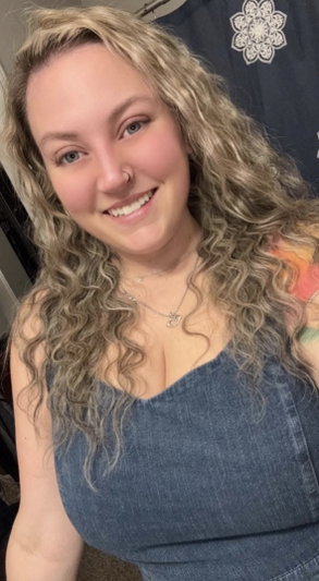 Woman with curly blonde hair wearing a denim top, smiling in an indoor setting.