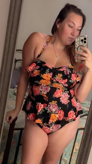 Woman in floral one-piece swimsuit taking mirror selfie in bedroom.