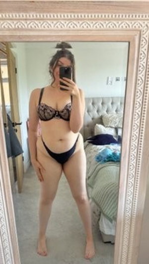 Woman in black lace bra and underwear takes mirror selfie in bedroom.