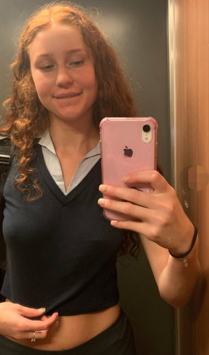 Woman with curly red hair in a dark crop top and black pants taking a mirror selfie indoors.