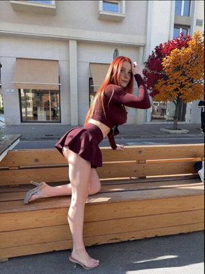 Woman with red hair in maroon outfit posing on wooden bench outdoors.