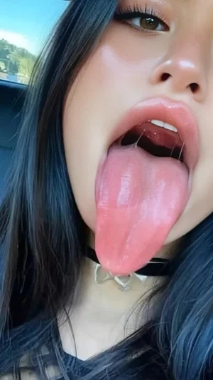 Woman with dark hair and choker sticking out tongue, indoors in car.
