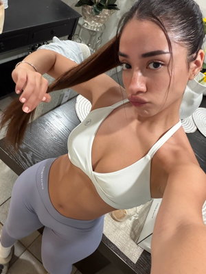 Woman in white sports bra and gray leggings posing indoors, pulling hair back.