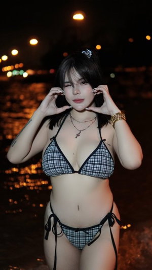 Curvy brunette in plaid bikini outdoors at night by water.