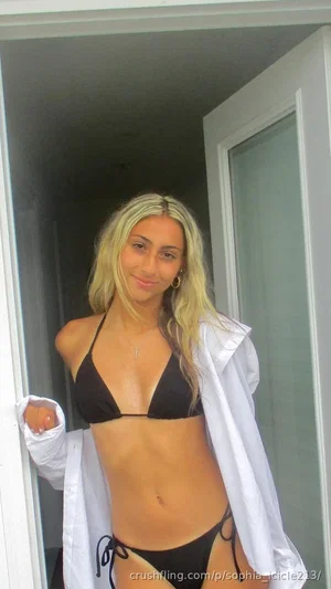 Woman with blonde hair in black bikini and white shirt standing in doorway.