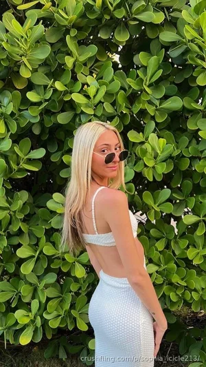 Blonde woman in white dress posing outdoors near green foliage.