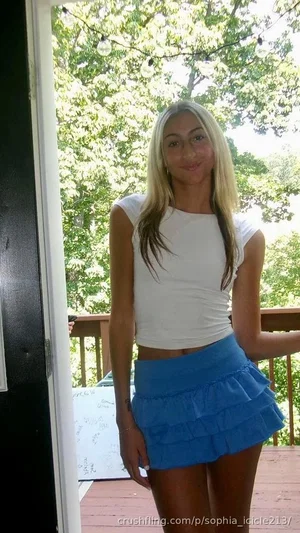 Woman with blonde hair in white crop top and blue ruffled skirt standing on wooden deck outdoors.