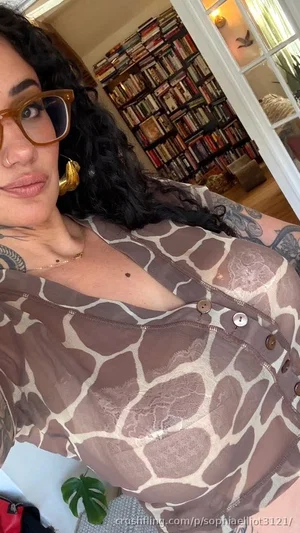 Woman with curly hair and glasses wears sheer giraffe-print top, indoors with bookshelf background.