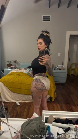 Woman with tattoos in black top and sheer shorts takes mirror selfie in bedroom.