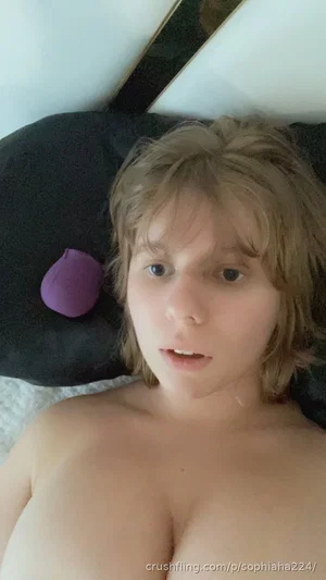 Woman with short blonde hair lying on bed, bare chest, black pillow with purple object nearby.