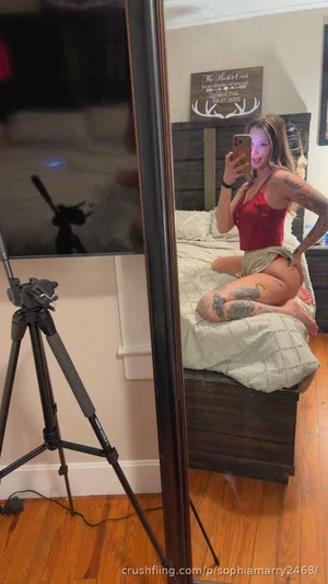 Woman with tattoos in red top and shorts sits on bed, taking mirror selfie in bedroom.