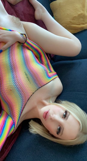 Blonde woman in rainbow mesh top lying on dark couch, hand on chest.