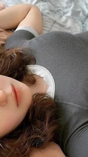 Woman with curly brown hair lying on bed, wearing gray top with white trim.
