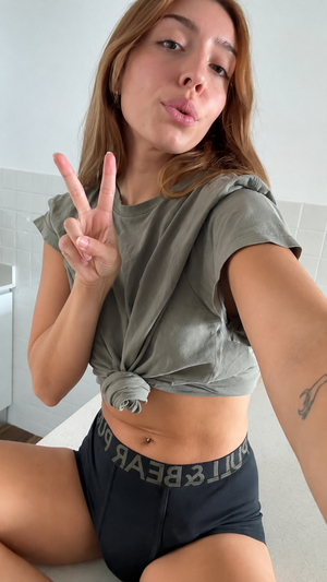Woman with light brown hair in olive crop top and black shorts, making peace sign indoors.