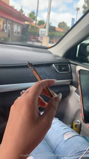 Person in car holding cigarette, wearing ripped jeans, hand with long nails visible.