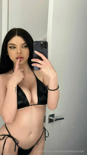 Woman with long dark hair in black bikini taking mirror selfie in bedroom.