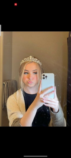 Woman with blonde hair wearing tiara and beige cardigan, taking mirror selfie indoors.