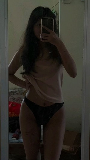 Slim Asian woman in beige top and black panties in bedroom mirror selfie.