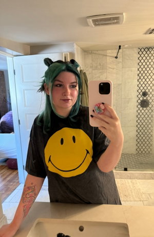 Woman with green hair in smiley shirt taking bathroom selfie.