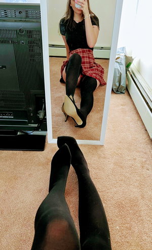 Woman in black top, plaid skirt, stockings, heels, mirror selfie.