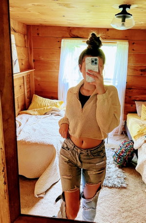 Woman with bun in cropped sweater, ripped shorts, bedroom mirror selfie.