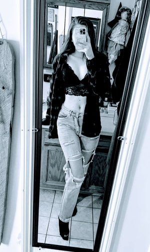 Woman in lace bralette, open plaid shirt, ripped jeans, bathroom mirror.