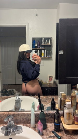 Woman in cap and hoodie taking mirror selfie in bathroom.