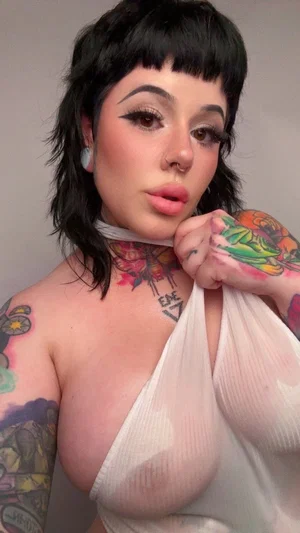 Woman with black hair and tattoos wears sheer white top, posing indoors.