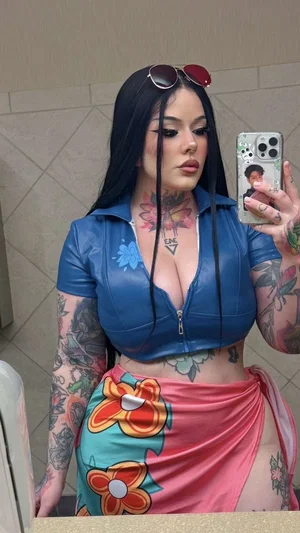 Woman with tattoos in blue crop top and floral skirt taking mirror selfie indoors.