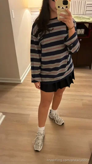 Woman in striped long-sleeve shirt and black skirt, standing indoors on wood floor, holding phone