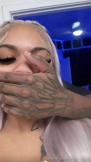 Woman with platinum hair and tattooed hand covering mouth, indoors.