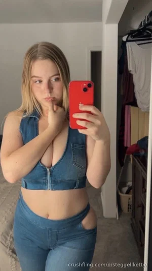 Woman in denim crop top and jeans takes mirror selfie in bedroom.