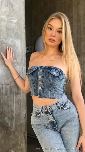 Blonde woman in denim crop top and jeans leaning against concrete pillar indoors.
