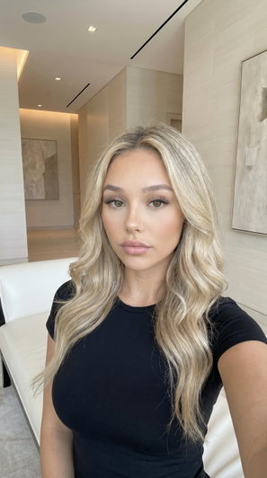 Woman with blonde wavy hair in black top, sitting indoors in modern room.