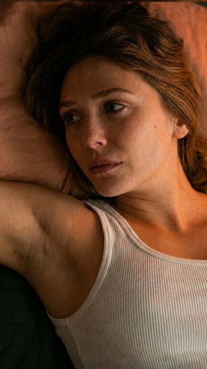 Woman with brown hair in white tank top reclining on bed.