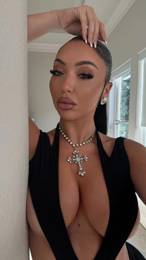 Woman in black plunging top with jewelry, posing indoors near a wall.