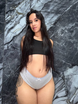 Woman with long dark hair in black crop top and gray underwear, standing against marble wall.