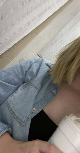 Blonde person in denim shirt and black top, holding cup, in tiled room.
