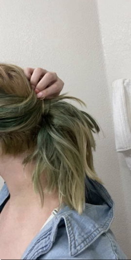 Person with green-tinted hair tying a messy bun, wearing denim jacket indoors.